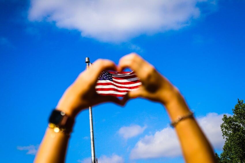 Two hands forming a heart shape with the American flag inside the formed shape and a blue sky